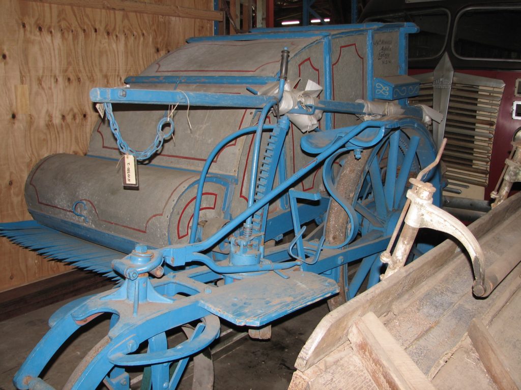 Coloured photograph of a harvester made of metal and finished with red lining and a blue frame. A comb at the front the crop drew it in towards enclosed beaters which removed the heads of wheat and forced it into a large box at the rear. The farmer sat on the harvester while holding the reins of the horses.